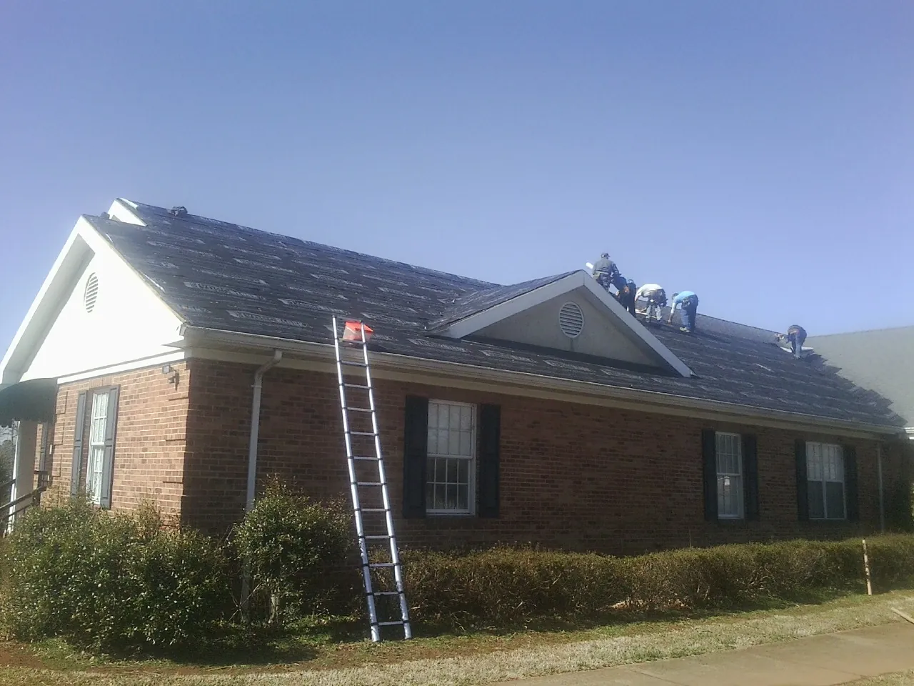 Crew tearing shingles off a brick ranch roof with a ladder set — Metal Roof Repair work by our crew in Alleman