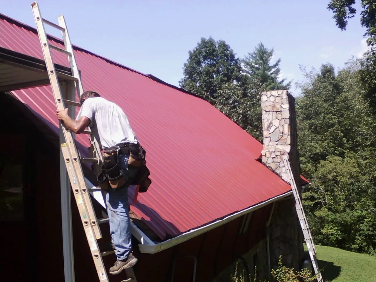 Roofer on a ladder beside a red metal roof and stone chimney — Soffit & Fascia Repair by Roofing Brothers in Alleman